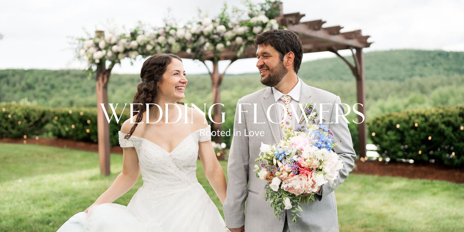 Two people walking hand-in-hand down a grassy pathway, surrounded by gardens and a an arbor, with the mountains visible in the background.