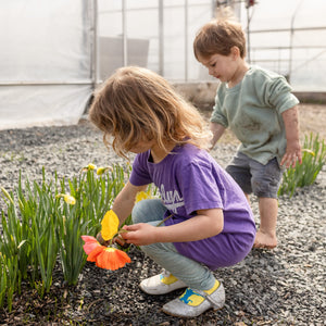 Two children in a greenhouse, one planting flowers and the other observing.