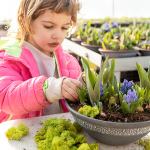 Child in pink jacket tending to plants in a pot with greenery on a table.