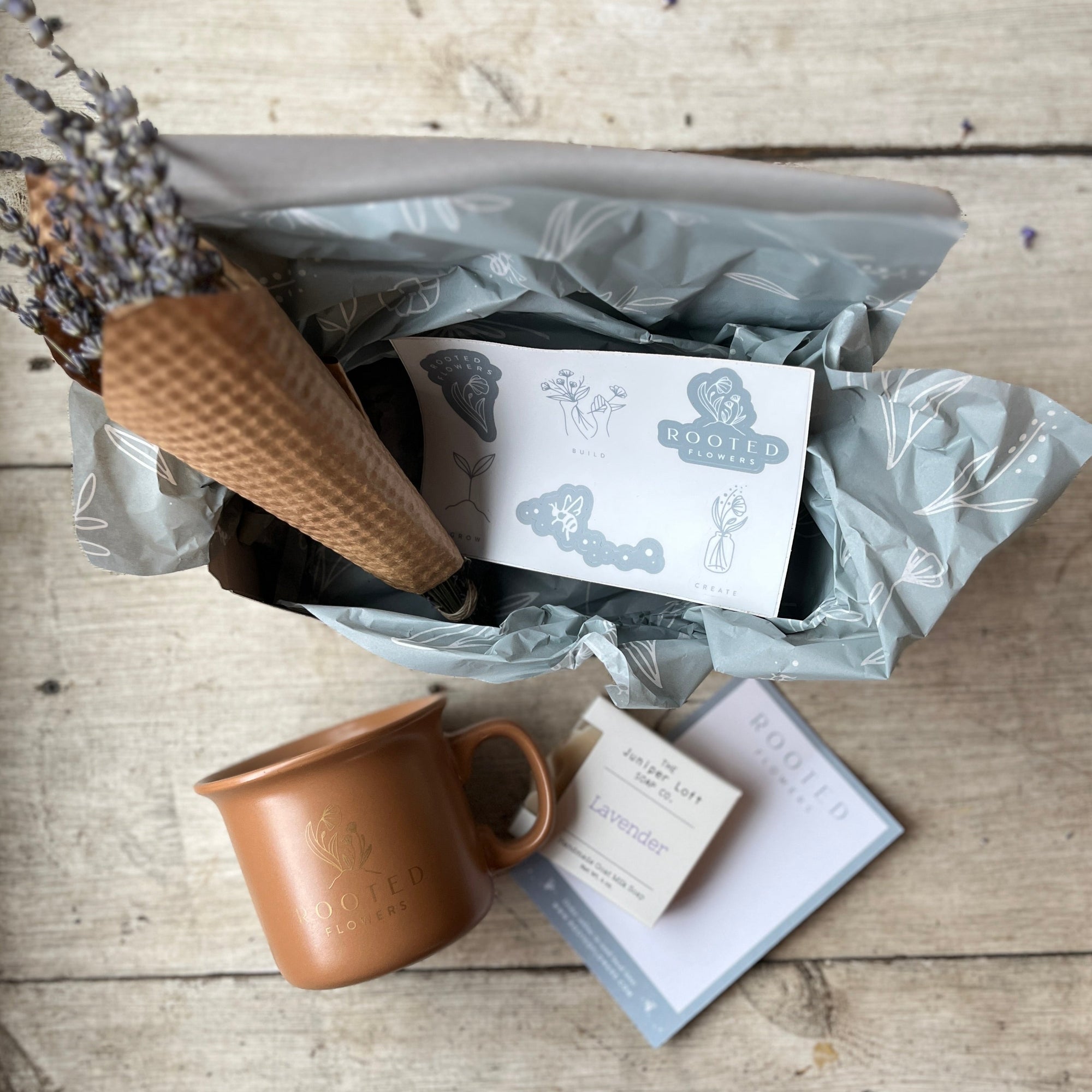 Image of the Lavender Bliss Box by Rooted Flowers, featuring an eco-friendly mug, a bunch of dried lavender, a bar of homemade lavender goat milk soap, and fun brand stickers, arranged neatly against a rustic wooden background. Perfect for back-to-school gifting.